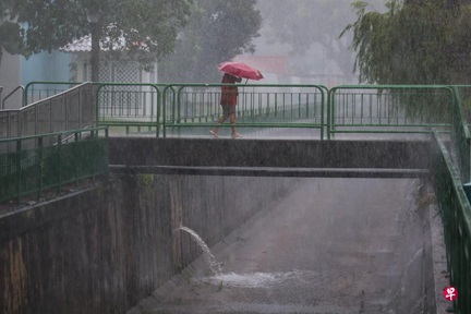 A person walks across a pedestrian bridge under heavy rain in Singapore, holding a red umbrella. Water is visibly gushing into a drainage canal below, highlighting the impact of intense rainfall on urban infrastructure.