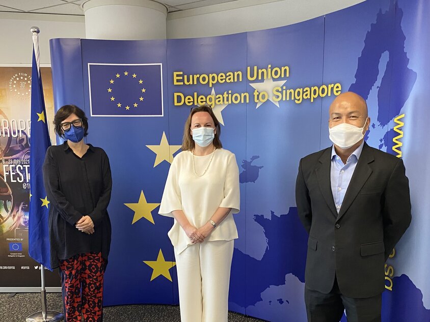 An indigo back drop with 3 people in masked standing before it.  On the left is a dark haired bespectacled western lady in black and patterned red pants, middle stands a lady in full cream top and pants, and on the right a bald man in suit without tie. 