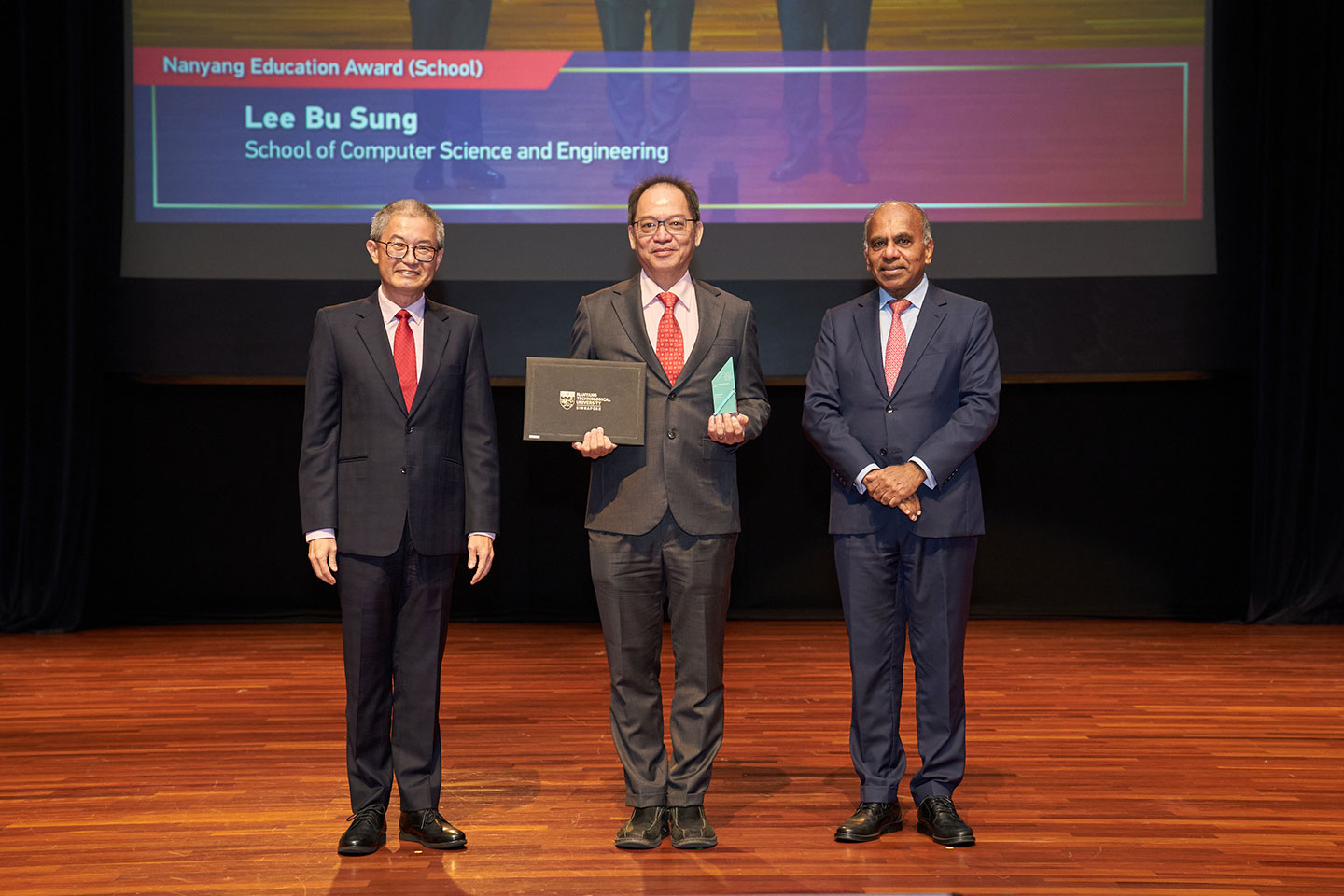 Photo of a professor standing between 2 presenters on stage holding a cert and an acrylic plaque.