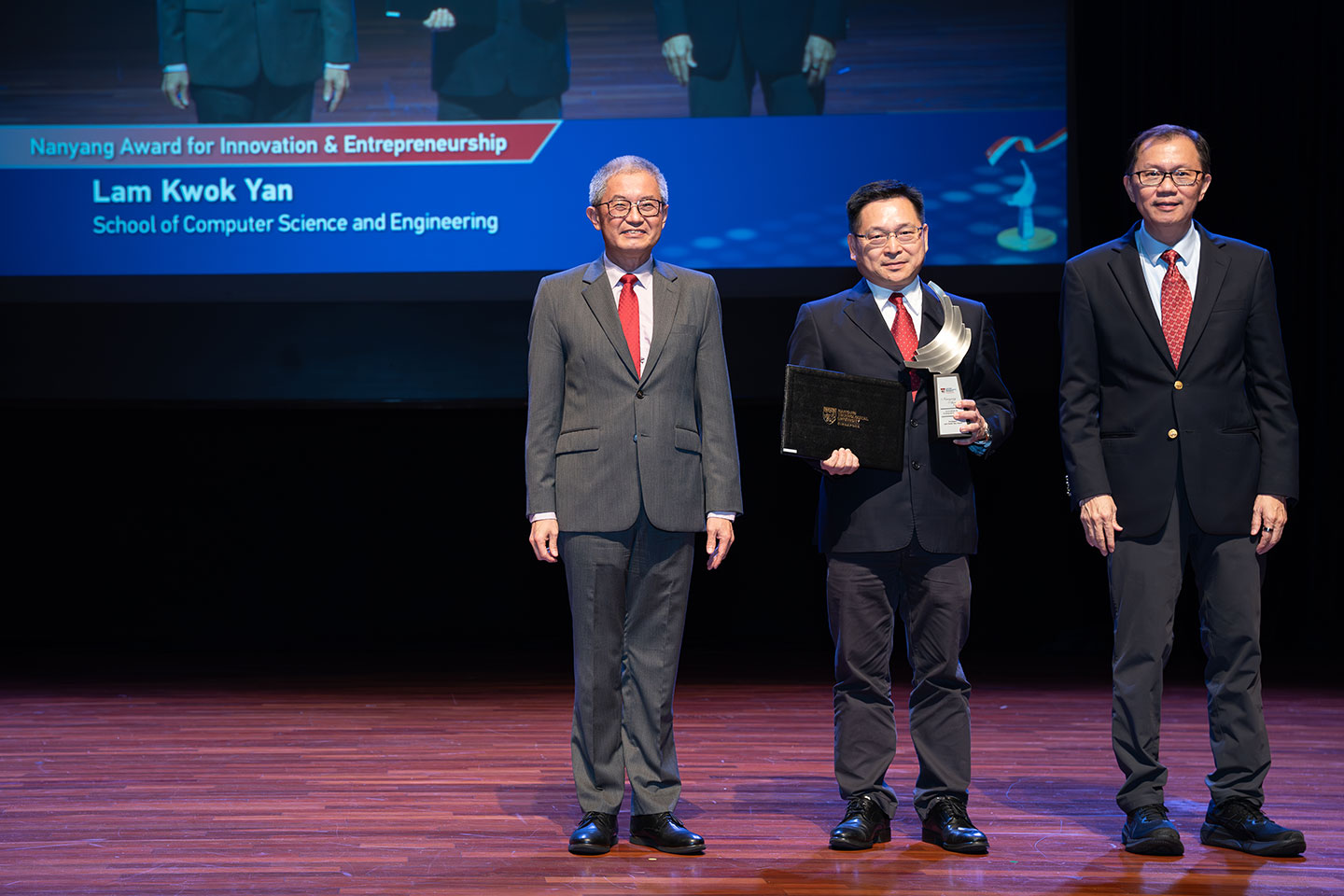 Photo of a Professor holding his award and certificate on stage with two Professors by his side on stage.
