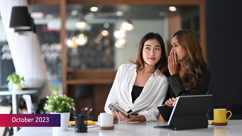 Woman whispering to a fellow female colleague with the text October 2023 on bottom left of image