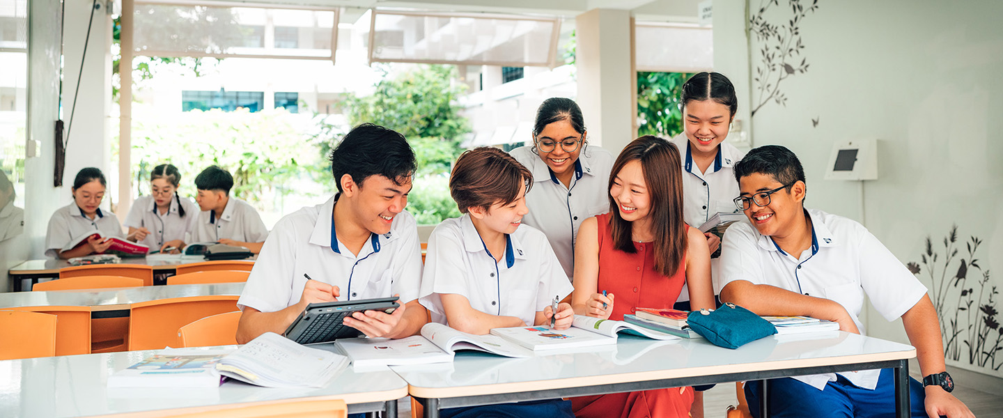 Female Teacher in Orange Dress with Group of 5 Secondary Students in Uniform Studying with Textbooks