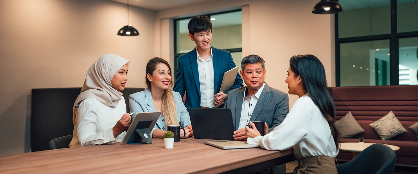 Older Male Teacher In A Grey Blazer With Group Of 4 Young Adult Students In Meeting Room