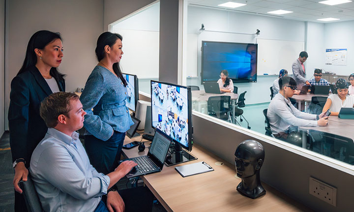 EEG Brain Caps SoLEC Classroom with Caucasian Man Looking At Computer Screen Monitoring Students Inside Of Classroom Behind The Glass Window (720x432px)