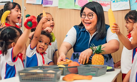 Pineapple and Toy Fruits Kindergarten Teacher and Students With Arms Raised (720x432px)