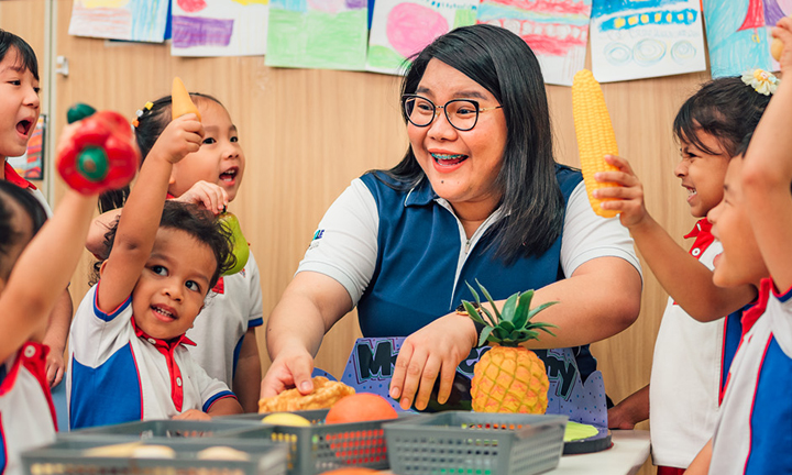 Pineapple and Toy Fruits Kindergarten Teacher and Students With Arms Raised 2 (720x432px)