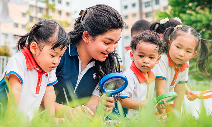 Toy Magnifying Glass Kindergarten Students And Teacher Outside On A Grassy Field Looking Down Inquisitively Closeup (720x432px)