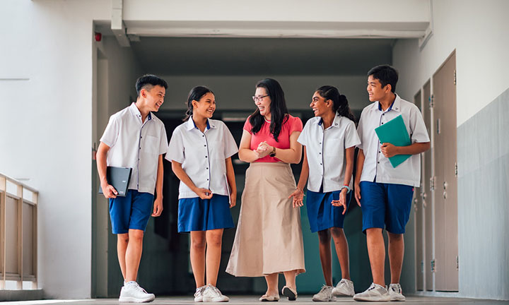 Walking Chinese Lady Teacher Red Top and Ivory Skirt Walking Along School Campus With Group Of 4 Secondary Students In School Uniform (720x432px)