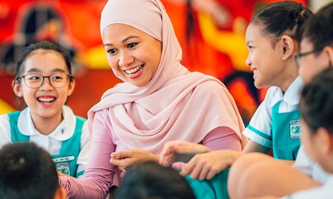 Yong Malay Lady Teacher With Pink Tudung With Group Of 8 Secondary School Students Smiling and Laughing Infront of Art Wall Closeup (720x432px)