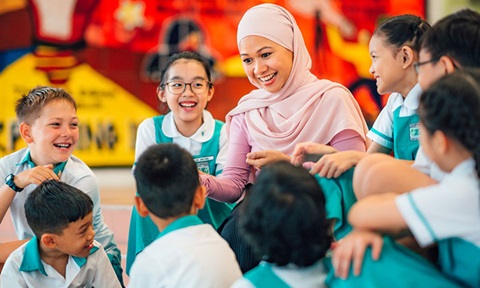 Yong Malay Lady Teacher With Pink Tudung With Group Of 8 Secondary School Students Smiling and Laughing Infront of Art Wall Wideshot (720x432px)