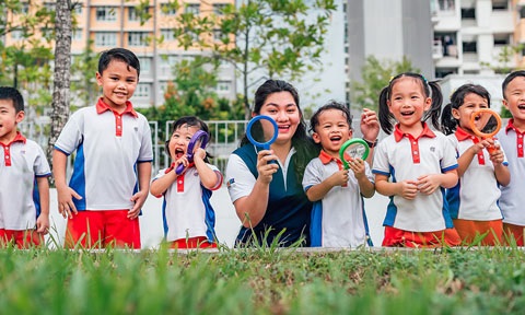 Toy Magnifying Glass Kindergarten Students And Teacher Outside On A Grassy Field Looking Straight Ahead All Smiles Wideshot (480x288px)