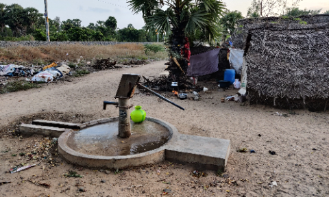 A hand pump in a rural setting surrounded by a small puddle of water and dry land, highlighting the challenges of water access and sanitation.