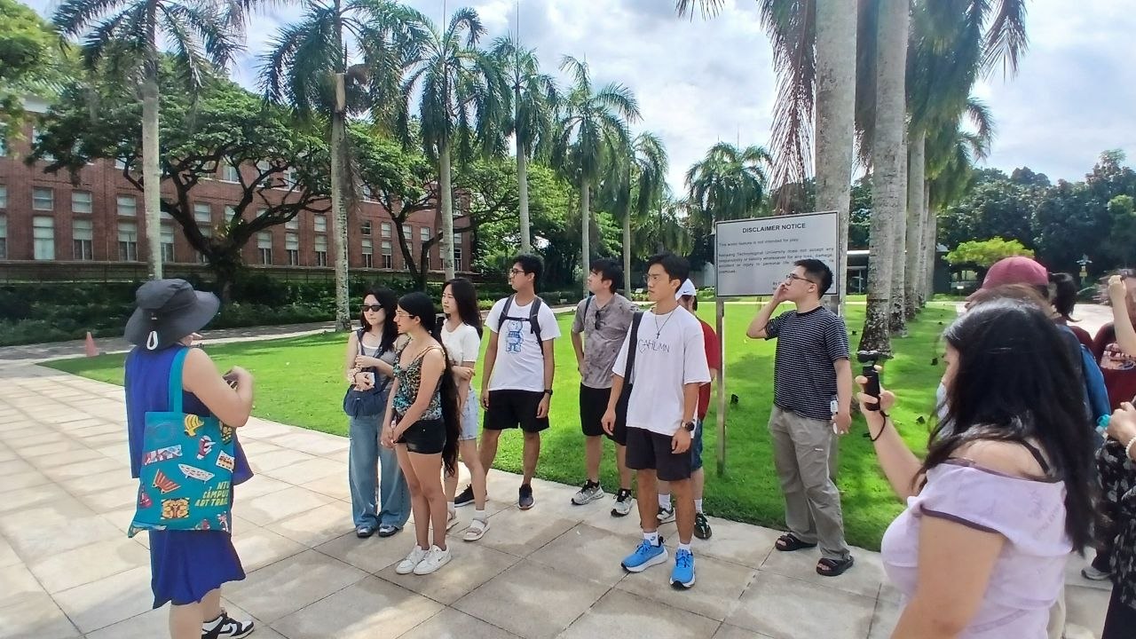 A group of people in front of a green lawn being addressed by a person in blue carrying a teal tote bag