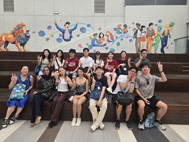 Group photo of people sitting on wooden steps in two rows in front of a colourful mural featuring a lion, happy people and graphic shapes