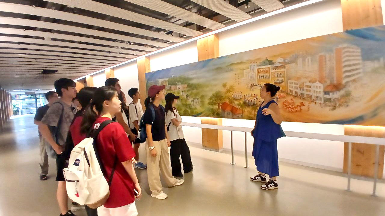 cat-walk-gem-summer-2025-group-wcy A group of people in a corridor looking at a long-handpainted mural, addressed by a person in blue