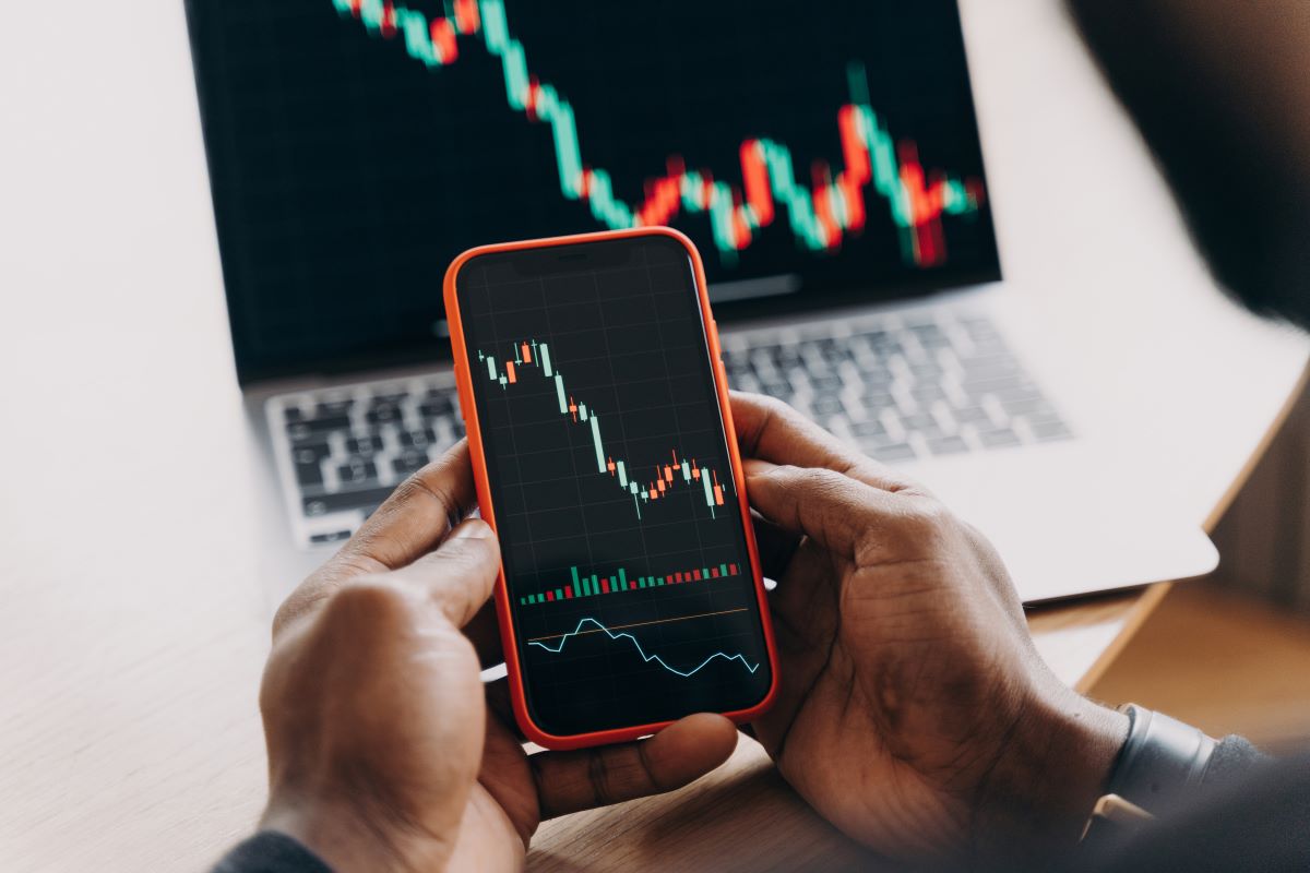 man stock trader holding mobile phone against laptop monitor