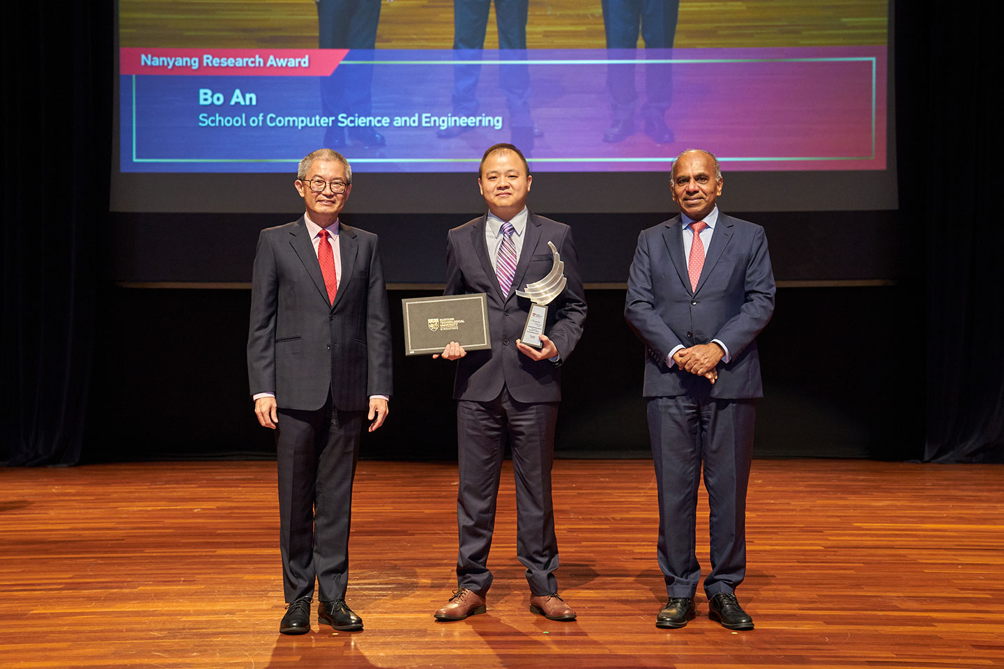 Photo of a professor standing in between 2 presenters on the stage, one hand holding a cert and the other hand holding a plaque.