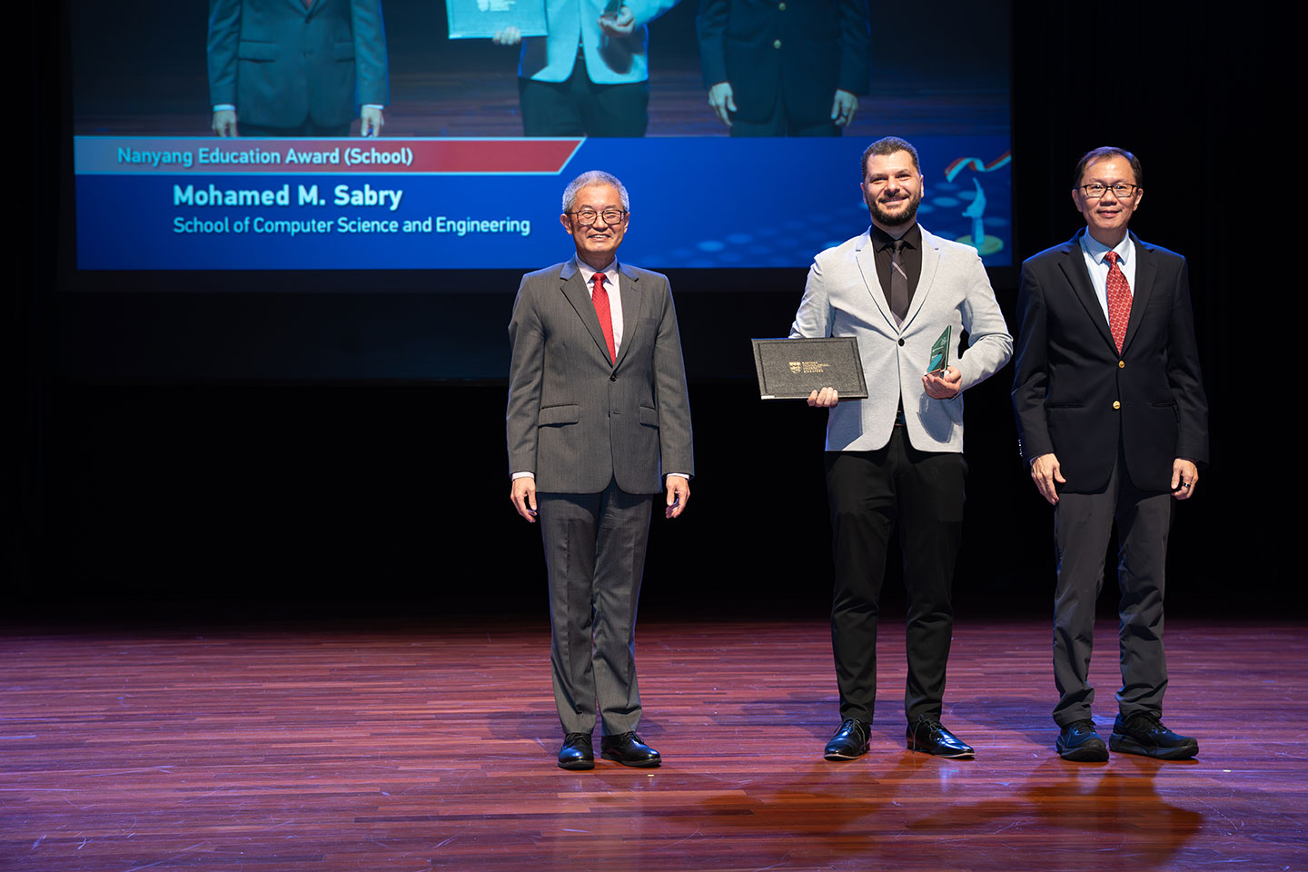 Photo of an Asst Professor holding his award and certificate on stage with two Professors by his side on stage.