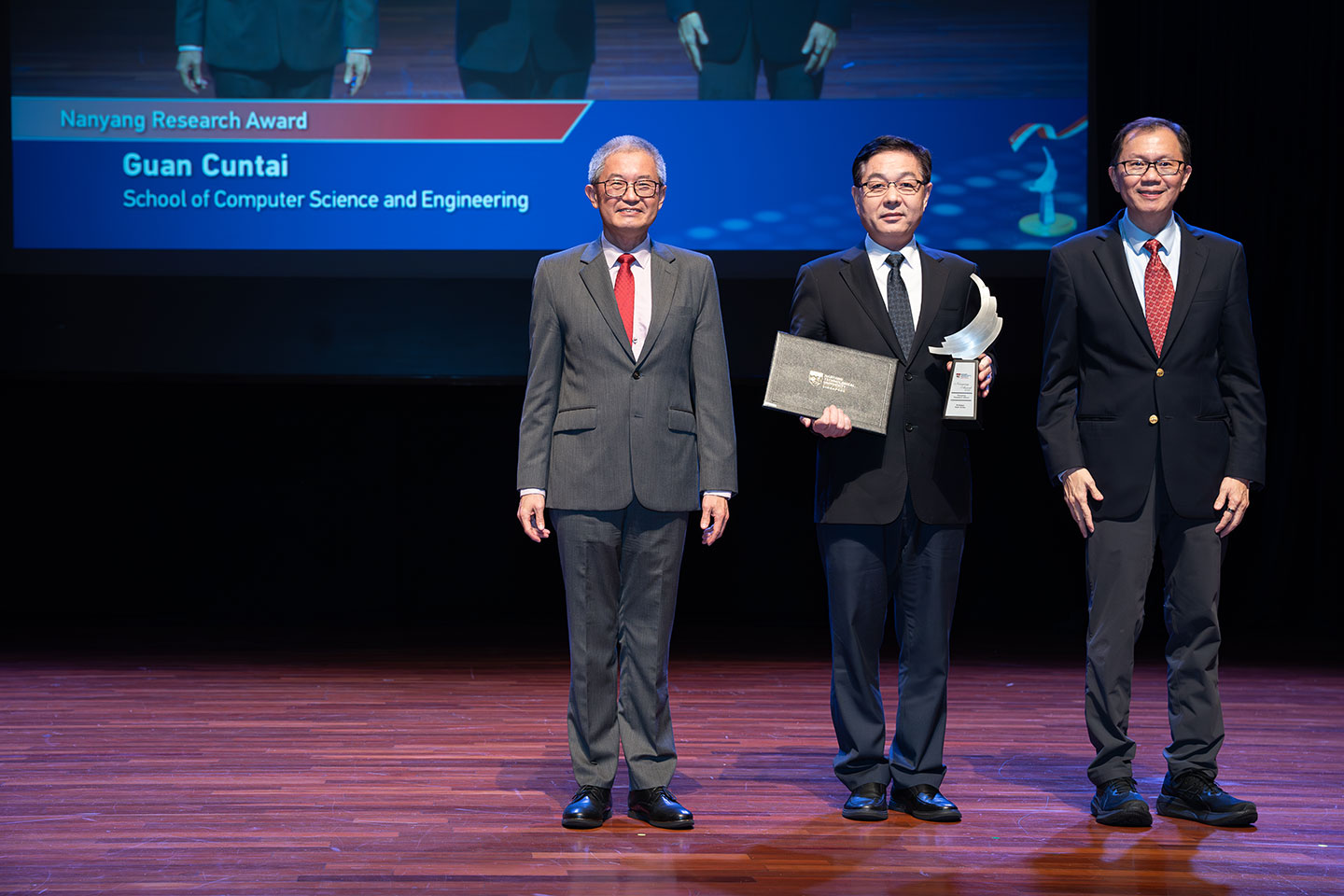 Photo of an Professor holding his award and certificate on stage with two Professors by his side on stage.