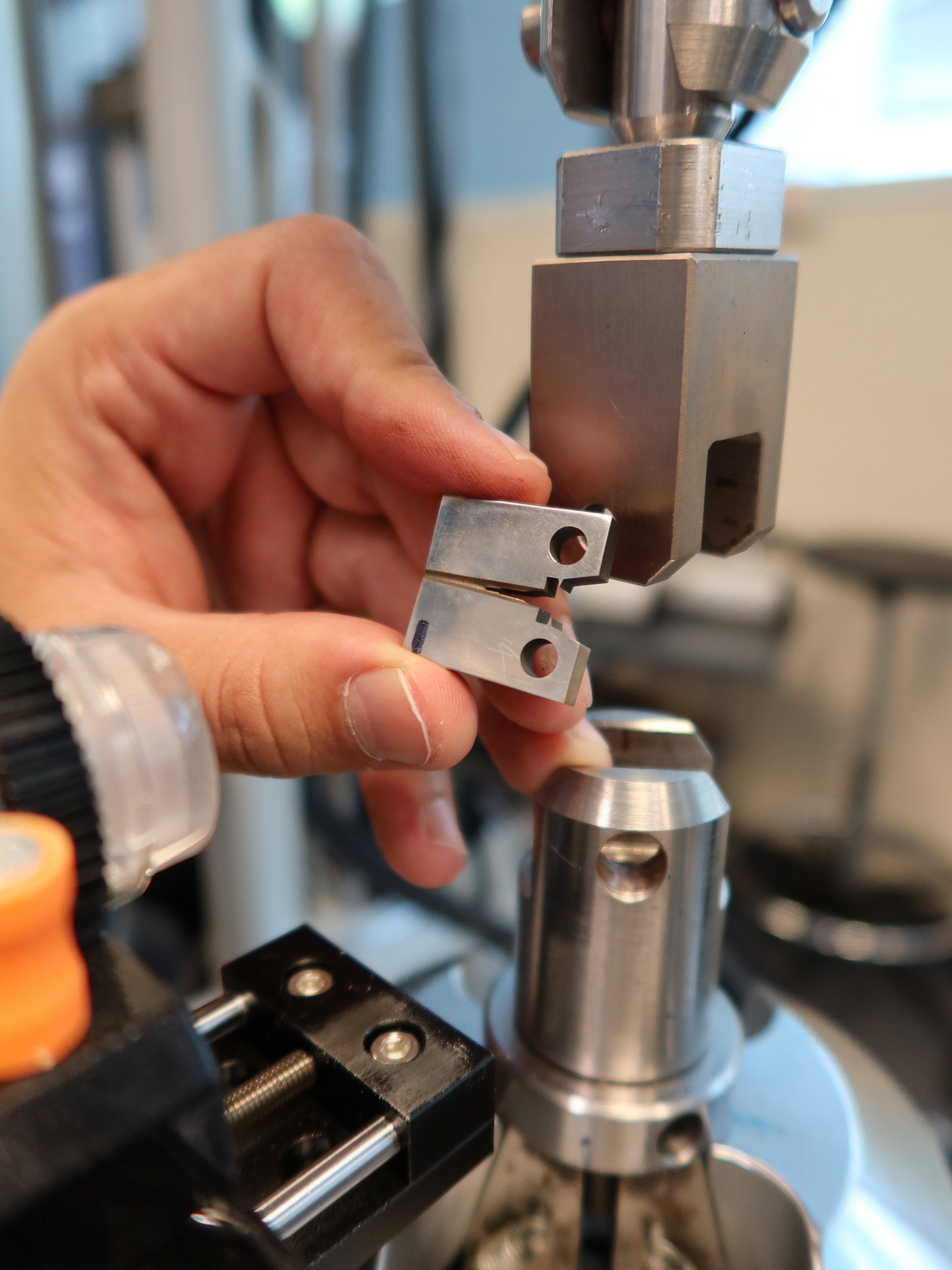 A hand holding a cracked metal test specimen beside a testing machine