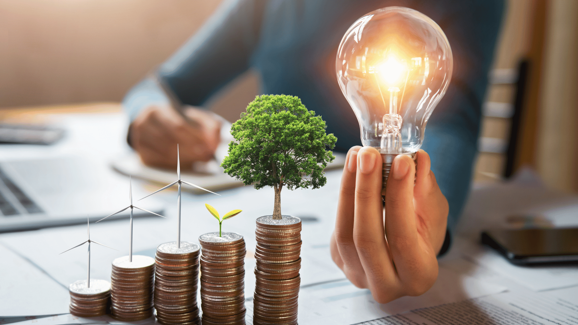 Stacks of coins with small plants and wind turbines beside hands