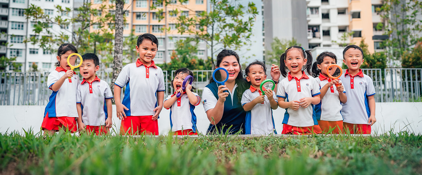 Toy Magnifying Glass Kindergarten Students And Teacher Outside On A Grassy Field Looking Straight Ahead All Smiles