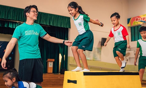 Male PE Teacher Instructing Group Of 4 Primary School Kids Jumping On Gymnastics Equipment Wideshot (720x432px)