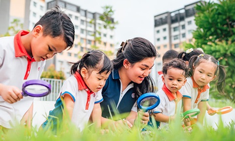 Toy Magnifying Glass Kindergarten Students And Teacher Outside On A Grassy Field Looking Down Inquisitively Wideshot (720x432px)