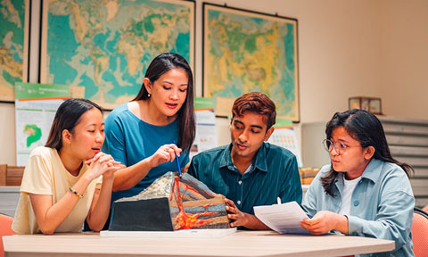 A Volcano Model Discussed by Group of 4 young students teacher blue dress wideshot (480x288px)