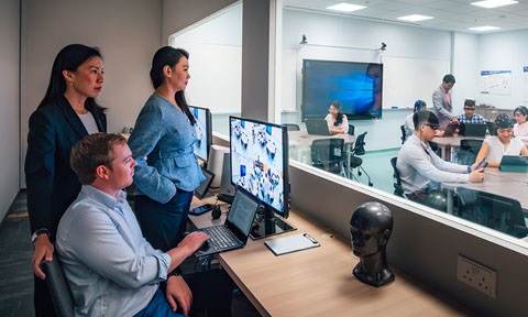 SoLEC Classroom with Caucasian Man Looking At Computer Screen Monitoring Students Wearing EEGs On The Inside Of Classroom Behind The Glass Window (480x288px)